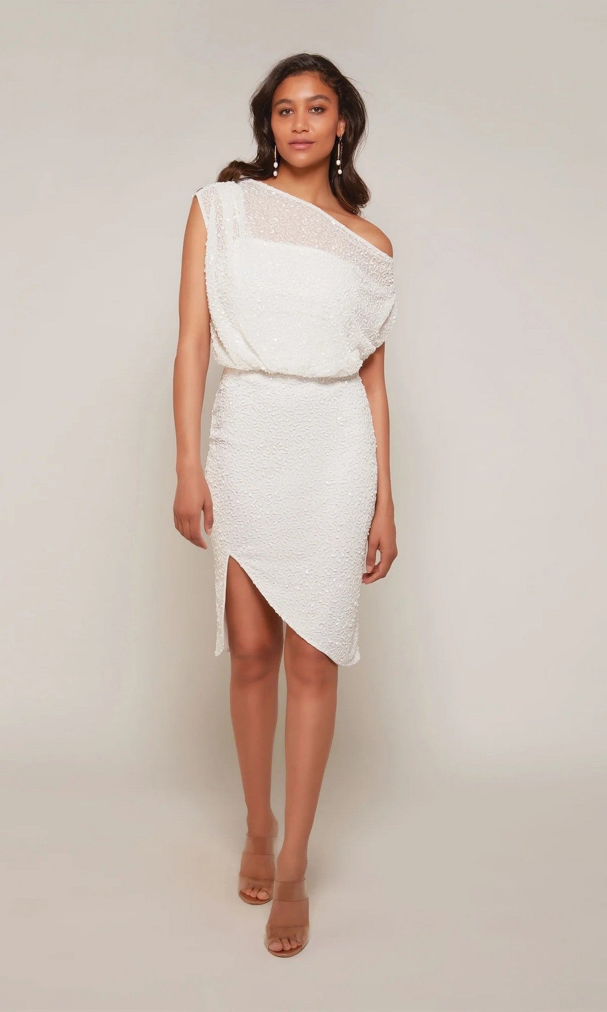 A woman stands against a plain background wearing the Alyce 70046 Knee Length White Party Dress, featuring an asymmetrical hem and one-shoulder neckline, styled with nude heels.